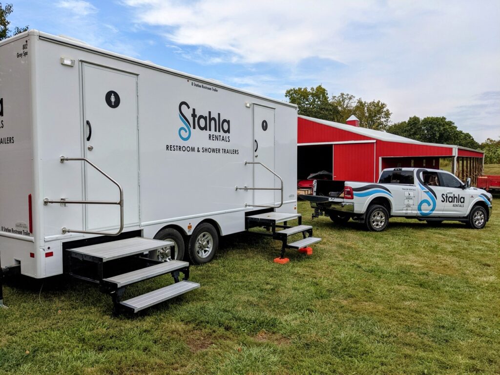a stahla rentals 8 stall restroom trailer rental is parked on the grass near a red building, with a pickup truck featuring the same company branding beside it.