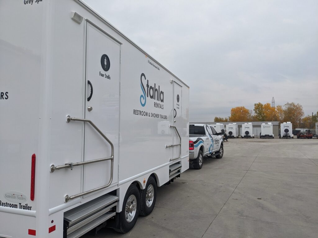 a row of 8 stall restroom trailer rentals and branded trucks lined up in an open lot under a cloudy sky. the trailers display "stahla rentals" logos.