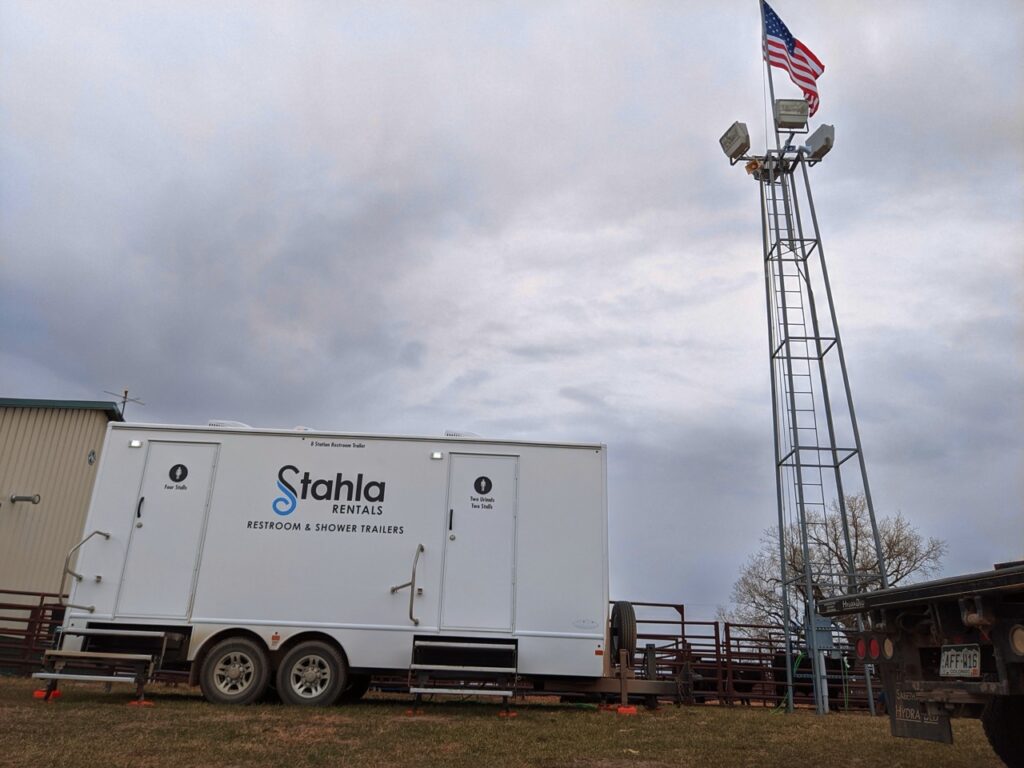 mobile 8 stall restroom trailer rental by stahla rentals parked on grass near a flagpole with the american flag and floodlights. a building and a parked truck are also visible.