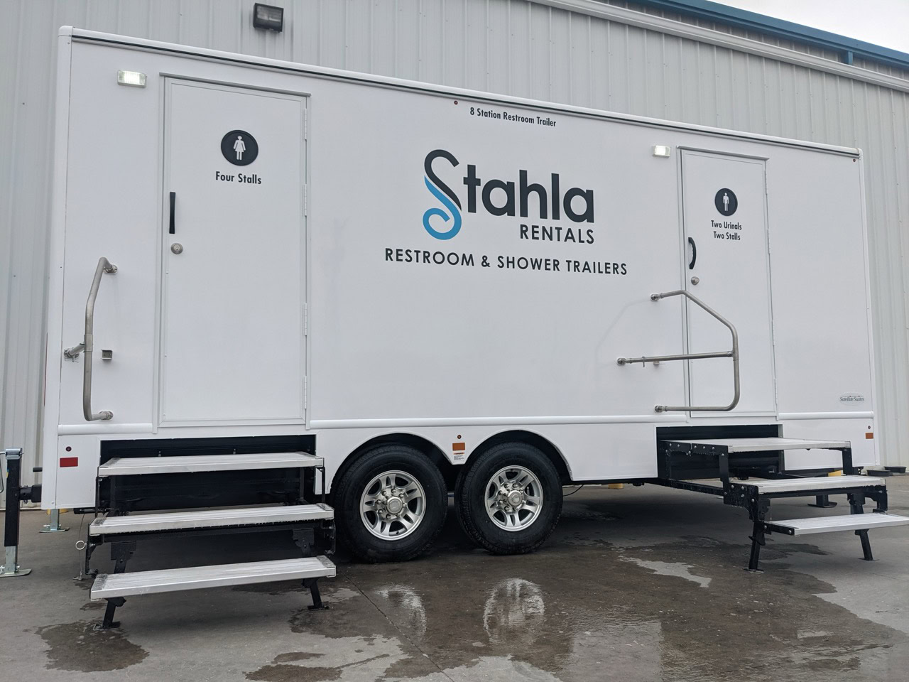 a white restroom and shower trailer with "stahla rentals" branding is parked outside. the 8 stall restroom trailer rental has two labeled doors: one for "four stalls" and another for "two stalls." two sets of stairs lead to the entrances.