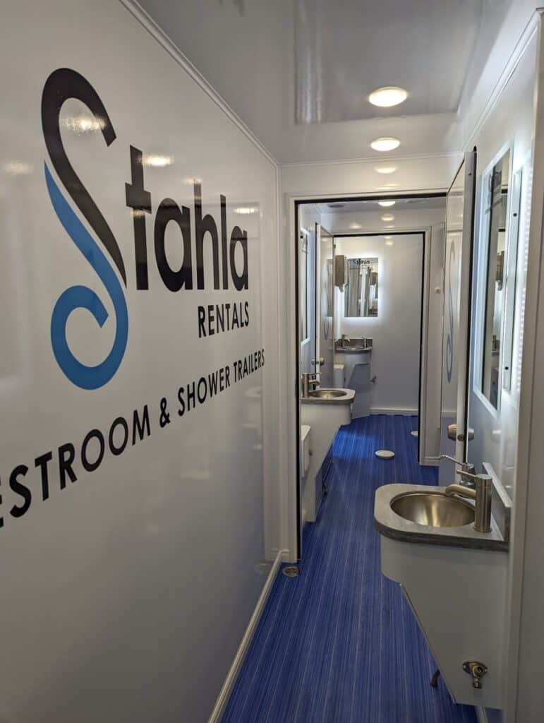 interior of a stahla rentals 8 station shower trailer, featuring sinks and mirrors along a corridor with blue carpeting.
