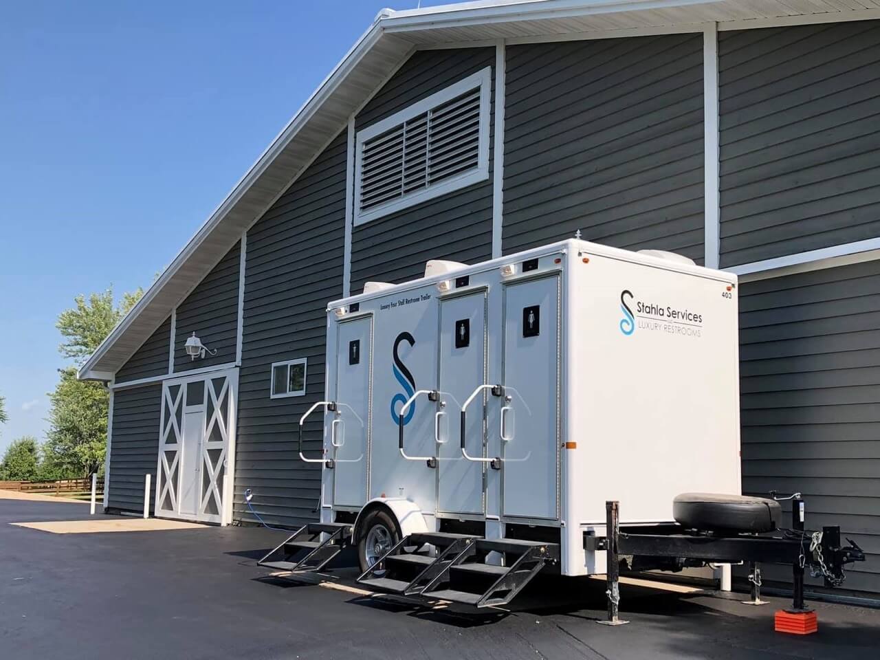 a 4 stall restroom trailer from stahla services is parked outside a large gray barn with white trim. the trailer features stairs and railings leading to three doors, offering convenient restroom access with a touch of elegance.