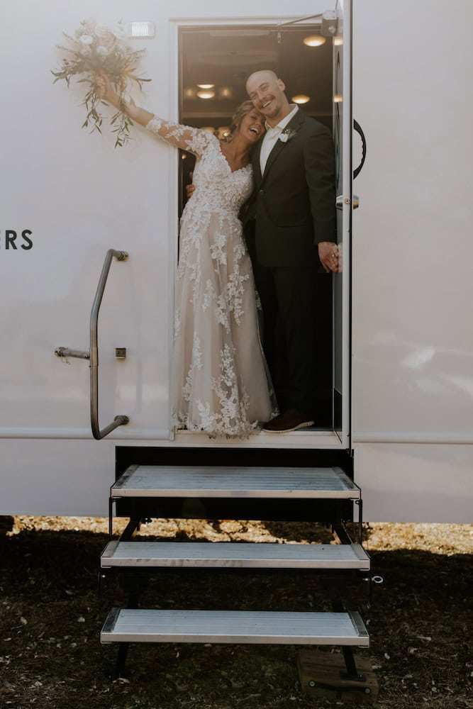 a joyful bride and groom pose in the doorway of a wedding bathroom rental trailer, the bride holding up a bouquet, both smiling warmly.