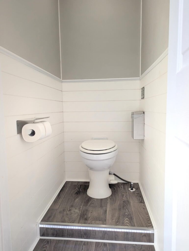 A Cozy Restroom Trailer Features A White Toilet, Wall Mounted Toilet Paper, Sleek Gray Wooden Floors, And Pristine White Panel Walls.