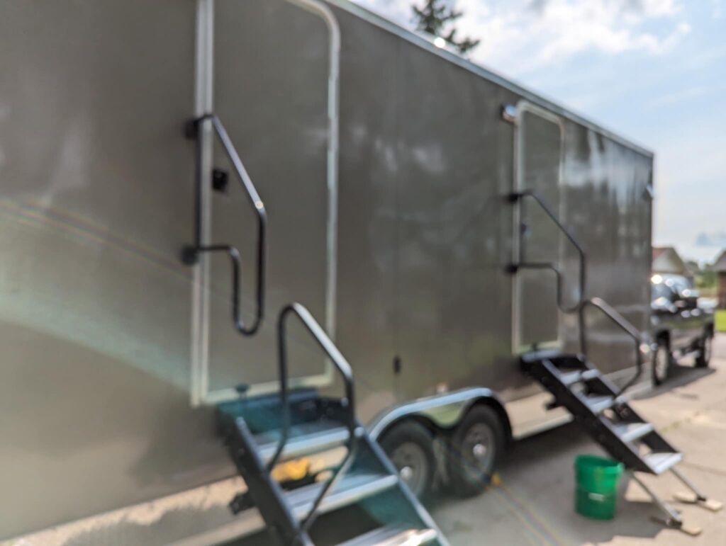 A Large, Gray Rt Trailer Is Parked On A Sunny Day, Featuring Multiple Steps And Doors. Beside It, A Green Bucket Adds A Pop Of Color To The Functional Landscape.