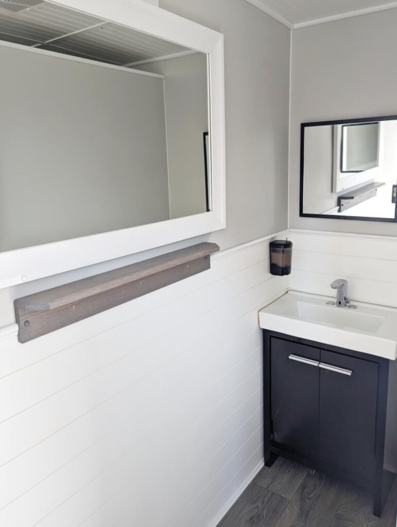 A Compact Restroom Trailer Features White Walls, A Rectangular Mirror, A White Sink With A Sleek Black Cabinet, And A Wall Mounted Toothbrush Holder.