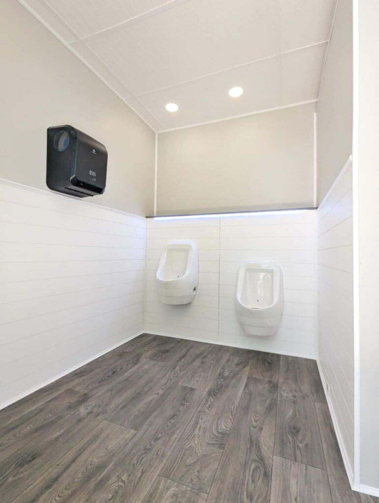 Two Urinals With A Dark Paper Towel Dispenser Are Mounted On A White Tiled Wall Inside The Restroom Trailer. The Room Features Wood Patterned Flooring And Is Softly Illuminated By Recessed Ceiling Lights, Showcasing The Attention To Detail Typical In Rt Trailers.