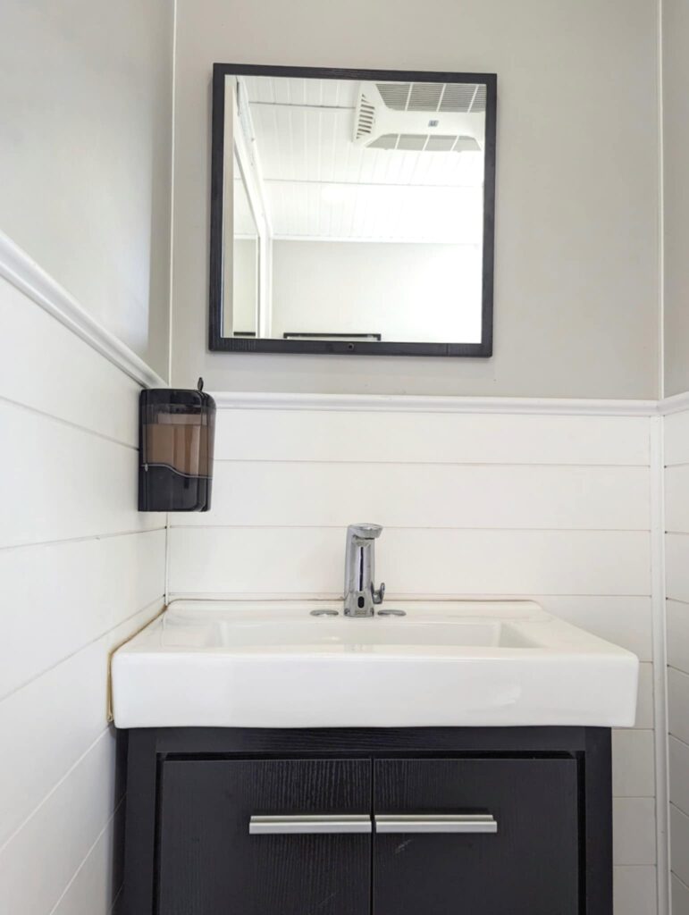 Small Bathroom With White Walls And Black Framed Mirror Above A White Sink And Black Cabinet. A Soap Dispenser Is Mounted On The Wall, Reminiscent Of Upscale Restroom Trailer Interiors.