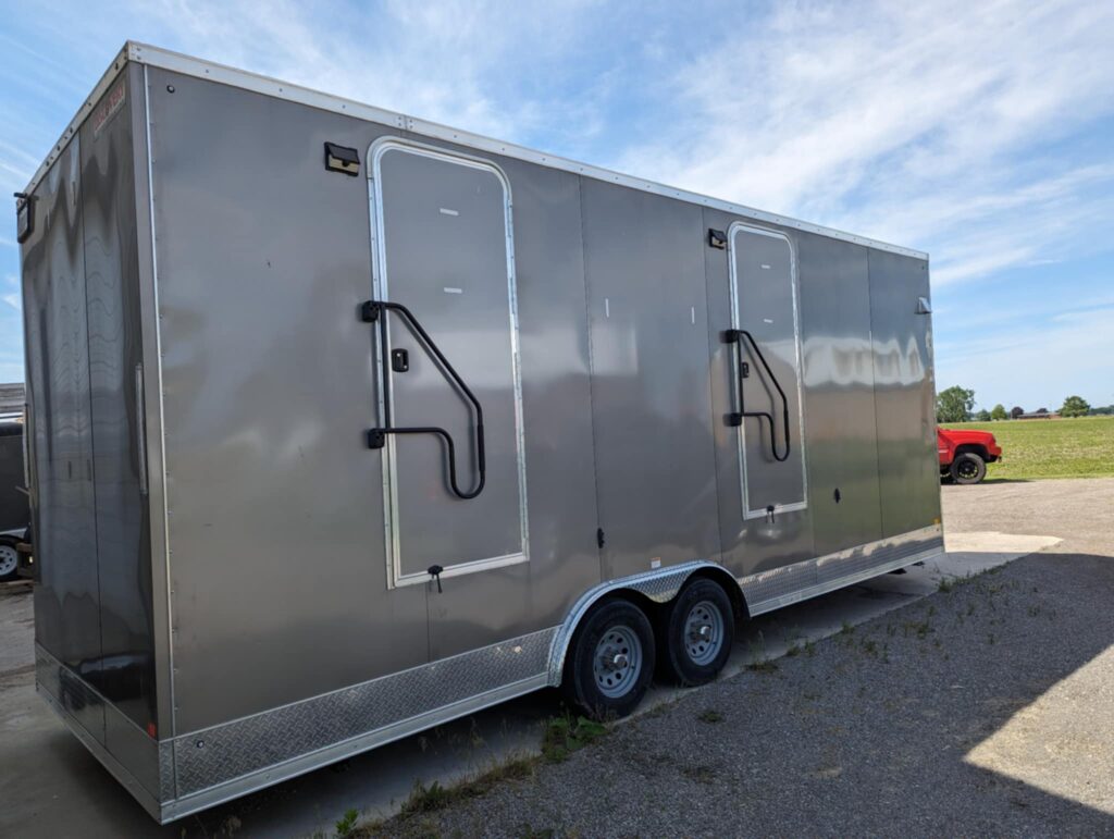 A Large Gray Restroom Trailer With Two Doors And Black Handles Is Parked On A Paved Area. A Red Vehicle And Fields Are Visible In The Background Under A Partly Cloudy Sky.
