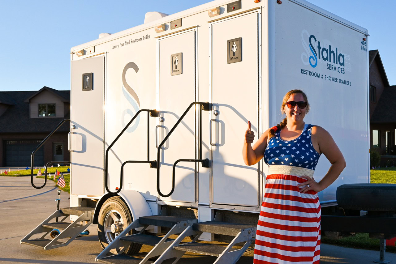 Woman In A Usa Flag Dress Gives A Thumbs Up In Front Of A Portable Restroom Trailer.