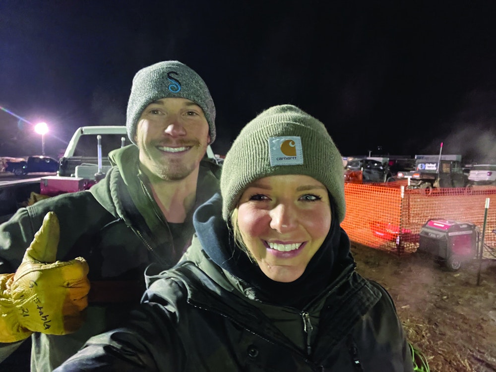 Two People In Winter Clothing Smile At The Camera. The Background Shows Vehicles And A Fenced Area, With Orange Netting And Lights.