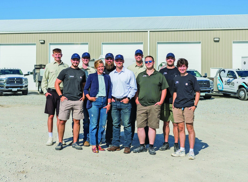 A Group Of Ten People, Including Six Men And Four Women, Stand Smiling In Front Of A Warehouse Building With Trucks Parked Nearby.