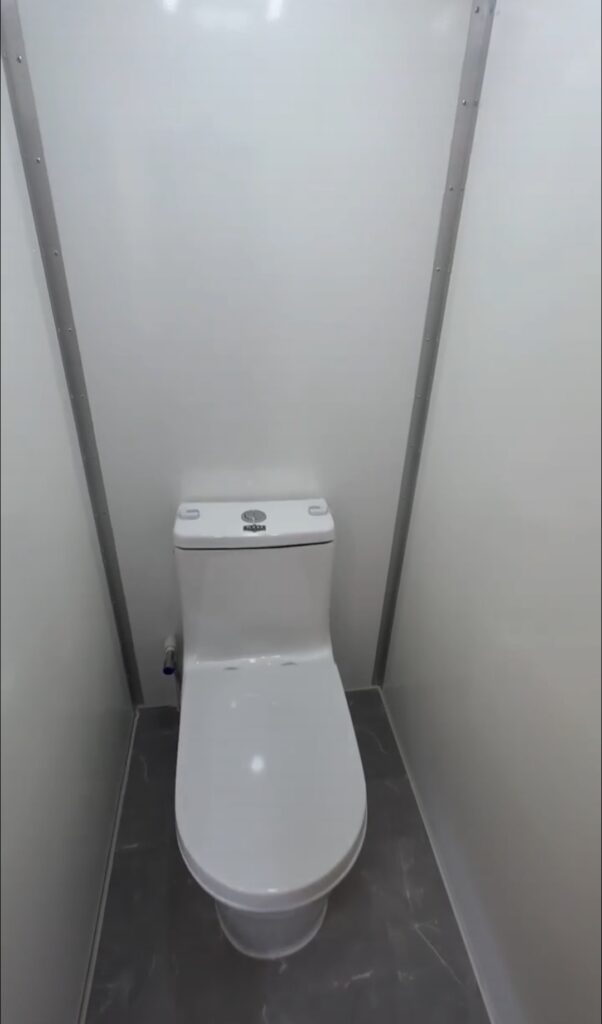 A White Toilet In A Small, Enclosed Stall With Gray Flooring And Light Colored Walls.