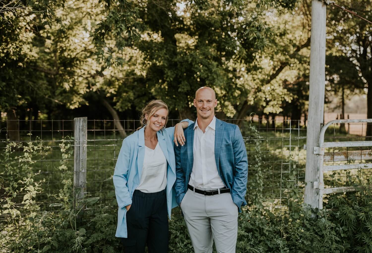 Two people stand side by side outdoors near a wire fence and gate, both wearing blazers and smiling, with greenery and trees in the background.
