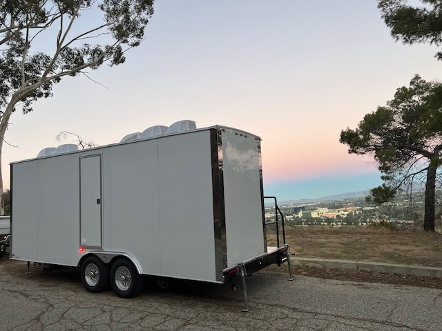 A white nuconcepts restroom trailer with rooftop vents is parked on the side of a road, overlooking a scenic valley at sunset.