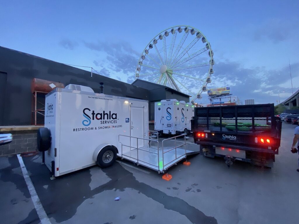 A stahla services restroom and shower trailer is parked next to a building, with a pickup truck and a large ferris wheel visible in the background.