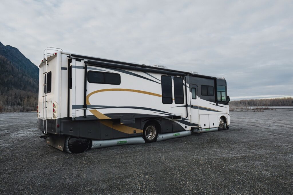 A large white rv with a gold and black stripe is parked on gravel with both rear tires visibly flat. trees and mountains are in the background under a cloudy sky.
