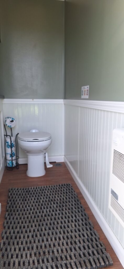 A small bathroom with a white toilet, toilet brush, patterned floor mat, green walls, and white wainscoting—perfect touches for a cozy 4 stall restroom trailer with a 4 season package.