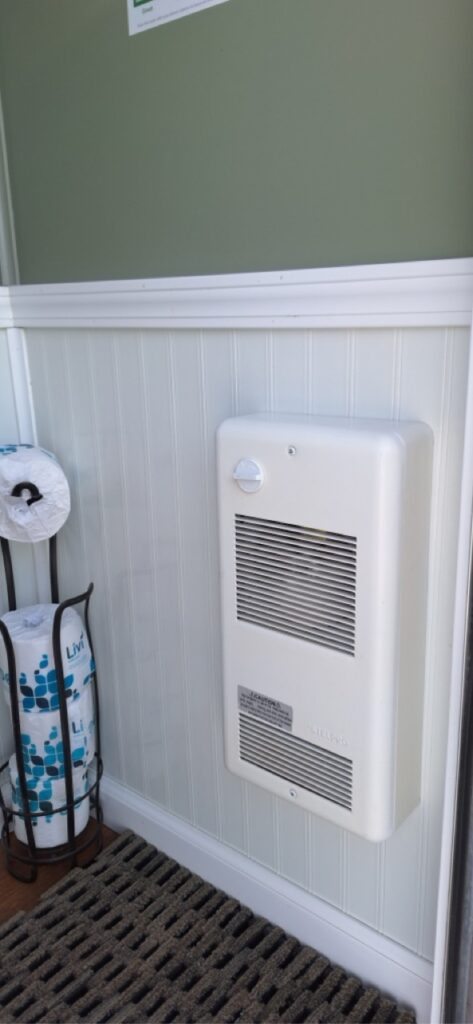 A wall mounted electric heater in a 4 stall restroom trailer is installed above a striped floor mat, next to a toilet paper holder with several rolls, against a green and white paneled wall.