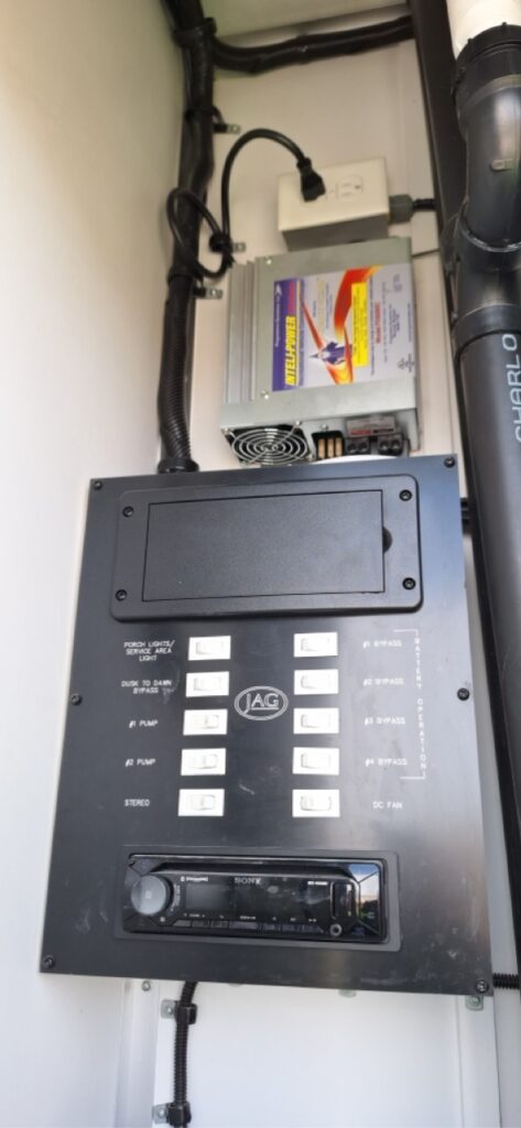 A control panel with labeled switches, a stereo unit, and an electronic power converter is mounted on a white wall with visible wiring, typically found in a restroom trailer for efficient operation.