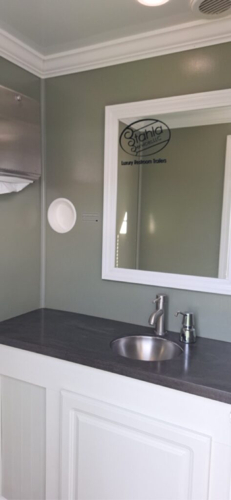 A small 4 stall restroom trailer with a gray countertop, stainless steel sink, mirror, and soap dispenser. The wall is green and the white cabinetry below the sink adds to this versatile 4 season package.