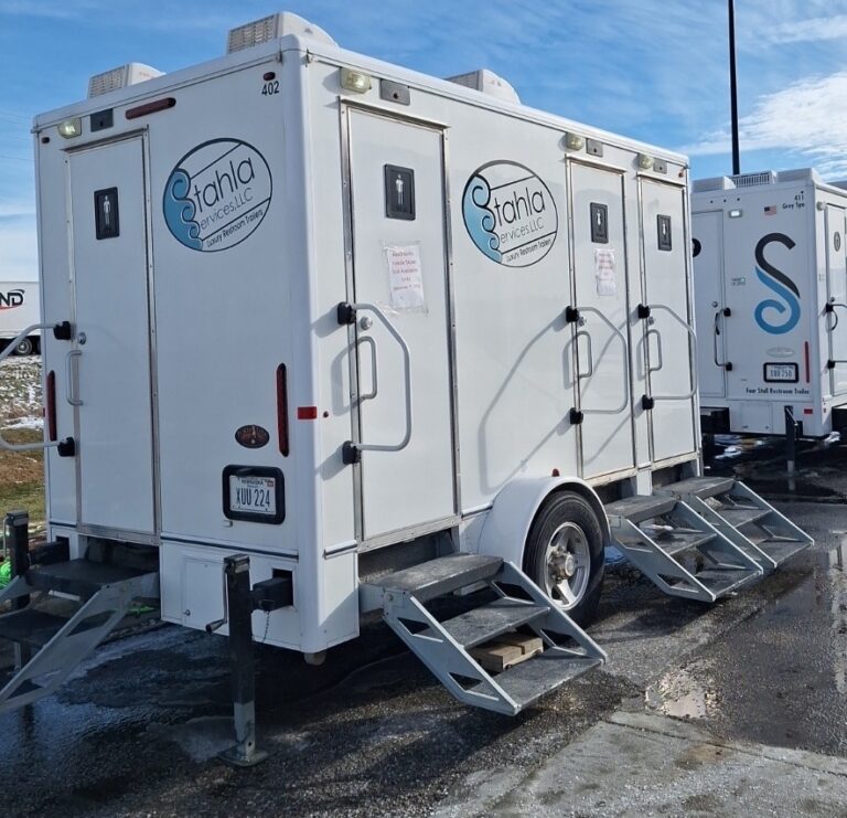 Two white 4 stall restroom trailers with metal steps are parked on a wet surface. Both trailers display the “Stahla Rentals” logo on their sides, showcasing their reliable restroom trailer designs.