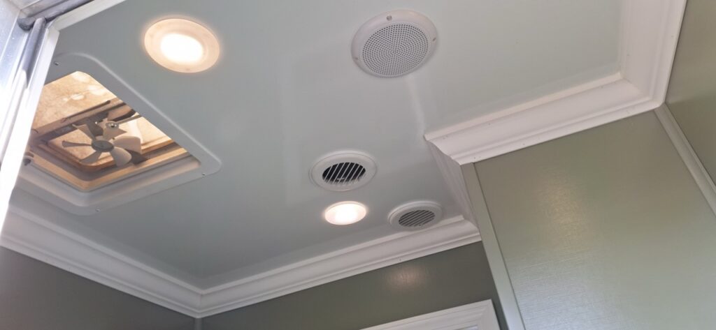 Ceiling of a 4 stall restroom trailer with recessed lights, circular air vents, a speaker, white crown molding, and an open ceiling vent fan—part of the versatile 4 season package.
