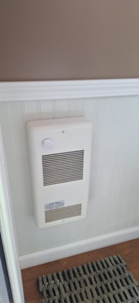 A wall mounted beige heater with a vent and control knob is installed on the white paneled lower wall beneath a brown upper wall, next to a striped doormat on a wood floor—ideal for 4 season comfort in a restroom trailer.