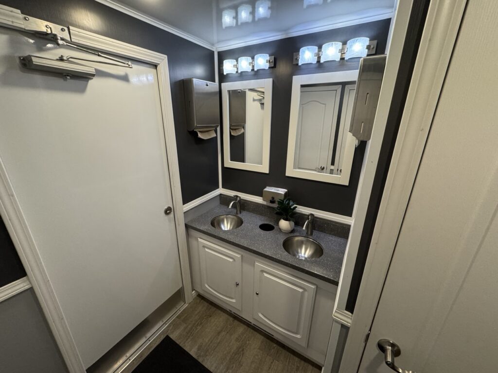 A modern 8 stall restroom trailer with two stainless steel sinks, a gray countertop, dual mirrors, bright vanity lights, a paper towel dispenser, and a decorative plant for a fresh touch.