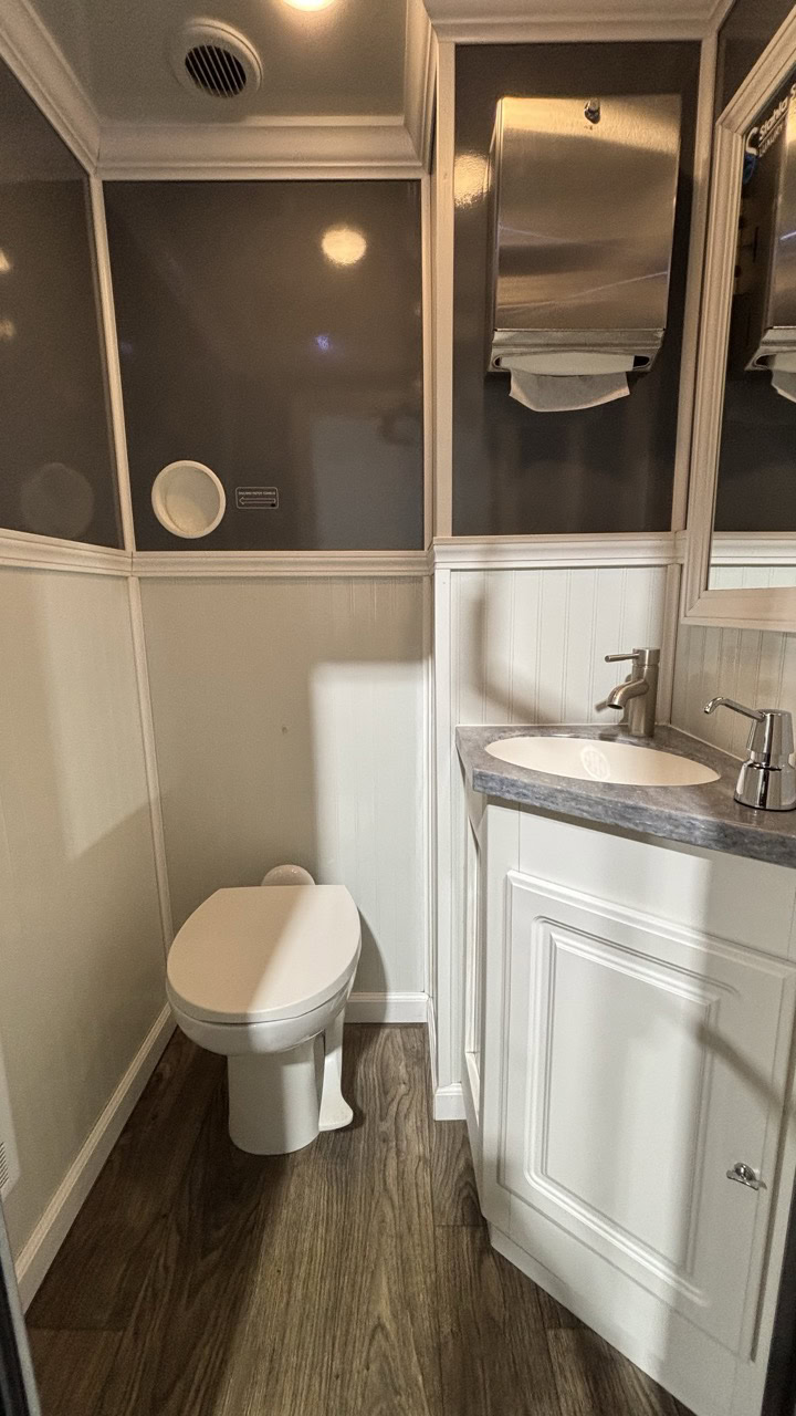 A small 4 Stall Restroom Trailer featuring a toilet, sink with cabinet, wall mounted paper towel dispenser, and wood style flooring.