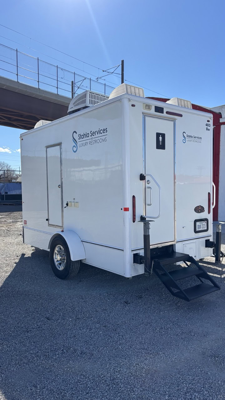 A white 4 stall restroom trailer labeled "Stahla Services Luxury Restrooms" is parked outdoors on gravel, featuring a 4 season package and stairs leading to the entrance.