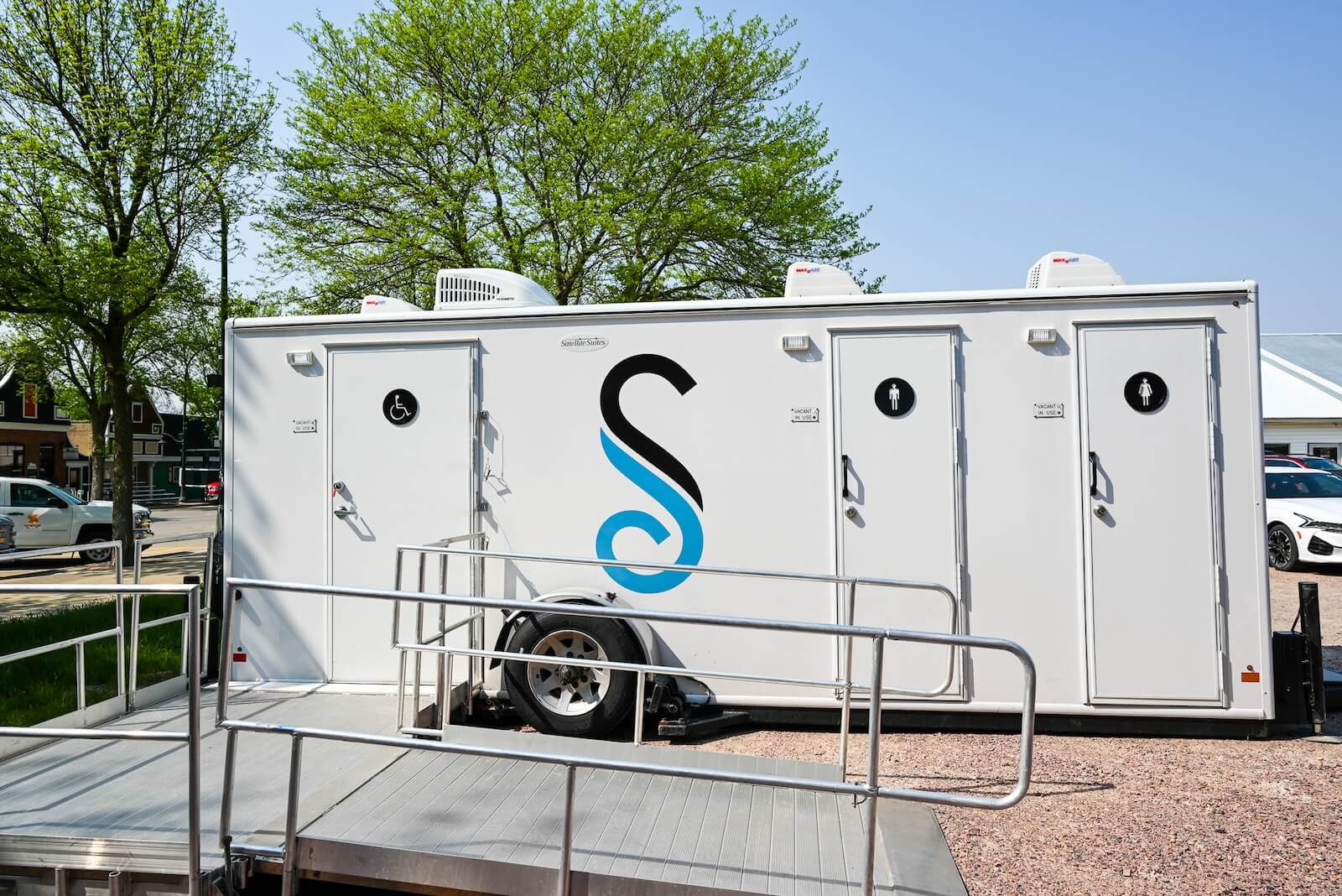 A white portable restroom trailer with three doors and a blue "S" logo is parked outdoors near a wheelchair accessible ramp, with trees in the background.