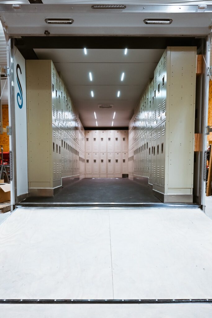 A view into a trailer outfitted with rows of beige metal lockers on both sides and the back, illuminated by ceiling lights.