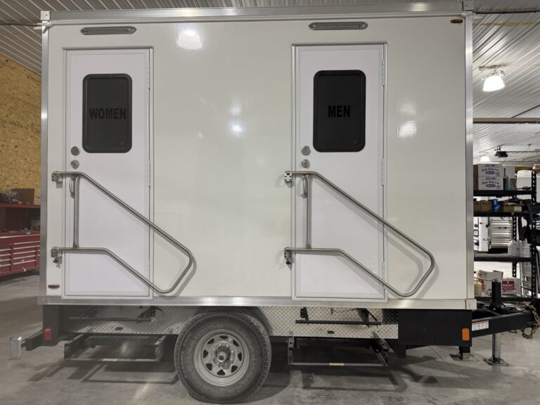 A 2 Stall Restroom Trailer with doors labeled "Women" and "Men," each featuring steps and handrails, is parked inside a garage or workshop—ideal for events in 2025.
